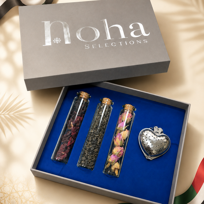 A luxury gift set from Noha Selections featuring three glass vials filled with dried botanicals and a silver heart-shaped teapot, presented in an open gray box with a blue interior, set against a warm beige background with ribbon accents.