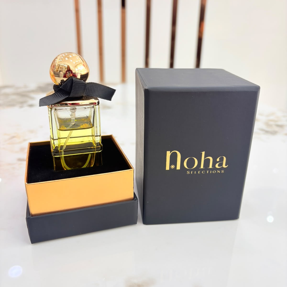 A bottle of Noha Scents perfume with a gold cap and black ribbon, displayed next to its dark gray and gold box on a light marble surface.