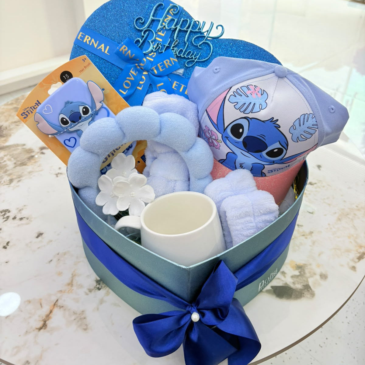 A blue and white Disney Stitch-themed baby gift basket set on a marble table, containing a plush toy, a teething ring, a small cup, and a card.