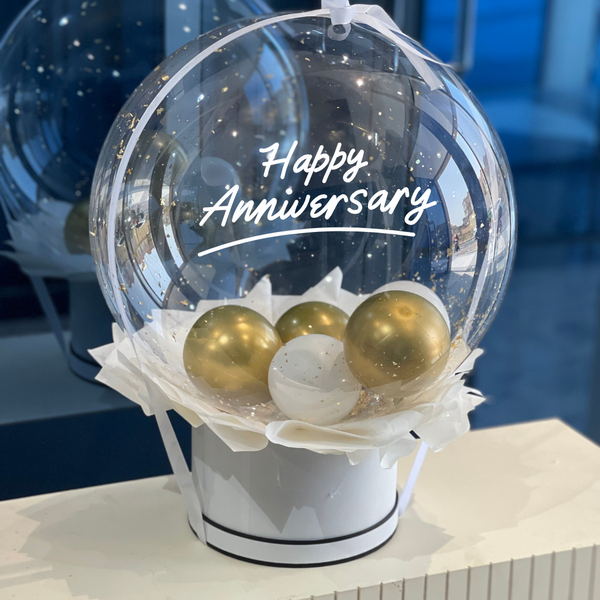 A transparent balloon with 'Happy Anniversary' written on it, containing smaller gold-colored balloons, presented in a white box with black trim.