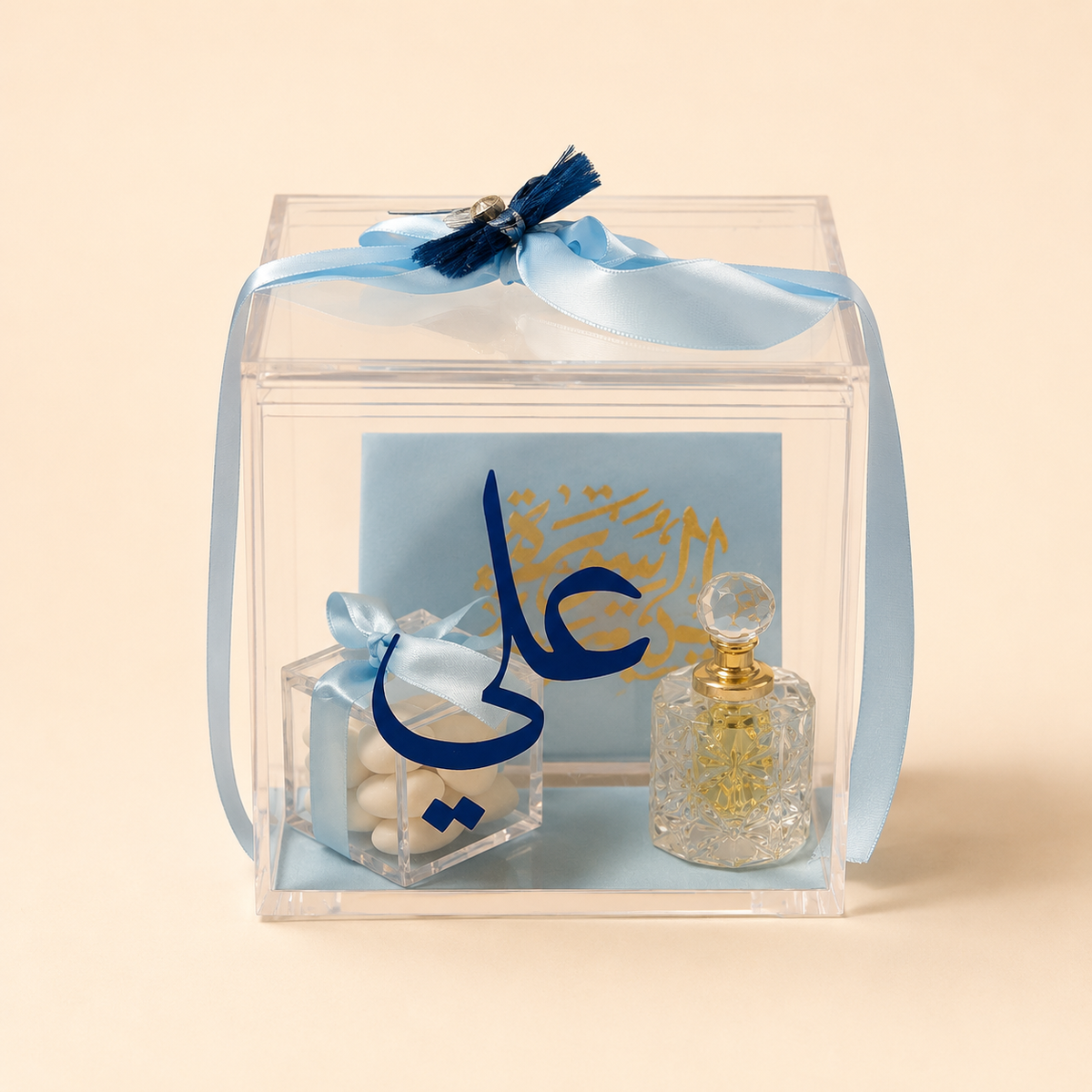 A clear acrylic gift box tied with a light blue ribbon, containing a small clear box of white candies with a blue ribbon, a blue card with gold Arabic calligraphy, and a small clear glass perfume bottle with a gold cap, set against a solid beige background.
