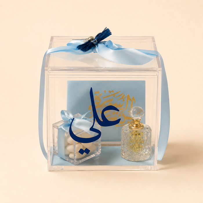 A clear acrylic gift box tied with a light blue ribbon, containing a small clear box of white candies with a blue ribbon, a blue card with gold Arabic calligraphy, and a small clear glass perfume bottle with a gold cap, set against a solid beige background.