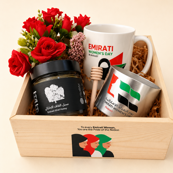 A curated gift basket for an Emirati woman, presented in a light wood box with red roses, a jar of Emirati Gold Honey, a white ceramic mug celebrating Women's Day, a wooden honey dipper, and a silver tumbler with the UAE flag.