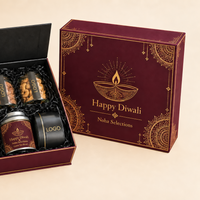 An open dark red and black gift box containing three small jars and a tin, displayed beside the matching closed box on a warm beige background. The closed box has gold decorative patterns and reads 'Happy Diwali Nisha Selections'.