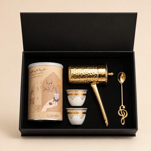 A luxury coffee gift set in an open black presentation box, featuring a gold-toned hand-drum coffee maker, two small white cups with gold patterns, a gold spoon, and a beige cylindrical tin canister with Arabic calligraphy.
