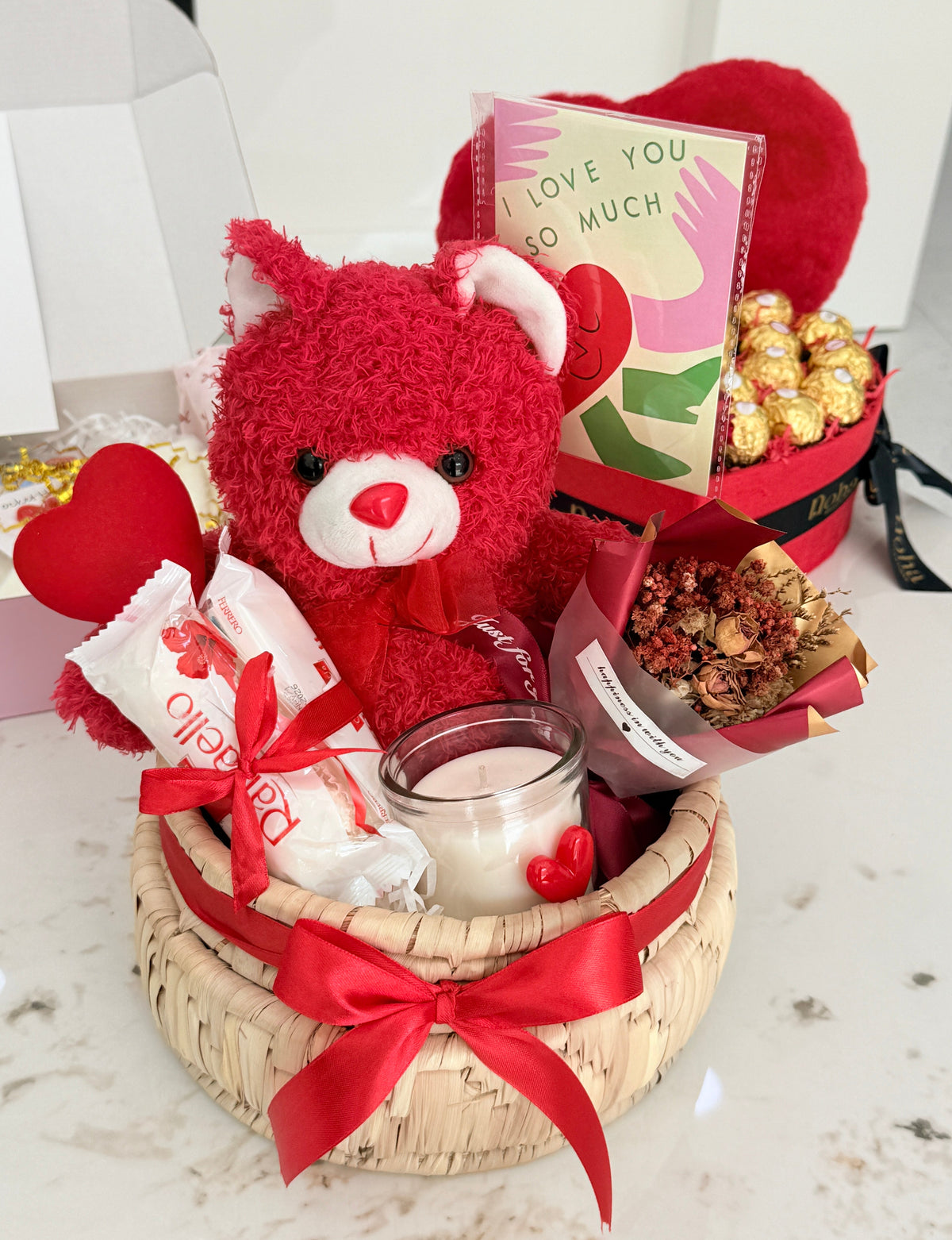 Gift basket with red teddy bear, chocolates, and a card on a white surface