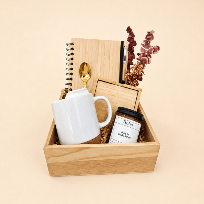 A curated gift basket in a light wood box featuring a white ceramic mug, a wooden notebook, a gold spoon, a small jar of palm leaves, and dried flowers, set against a solid beige background.