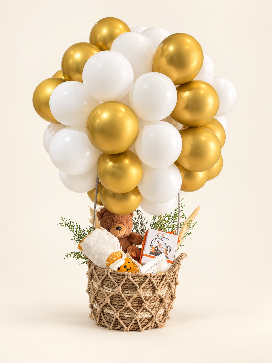 A hot air balloon-themed baby shower decoration set featuring a cluster of white and metallic gold balloons floating above a woven basket filled with baby toys, a plush teddy bear, a pacifier clip, and a small book, set against a solid cream background.