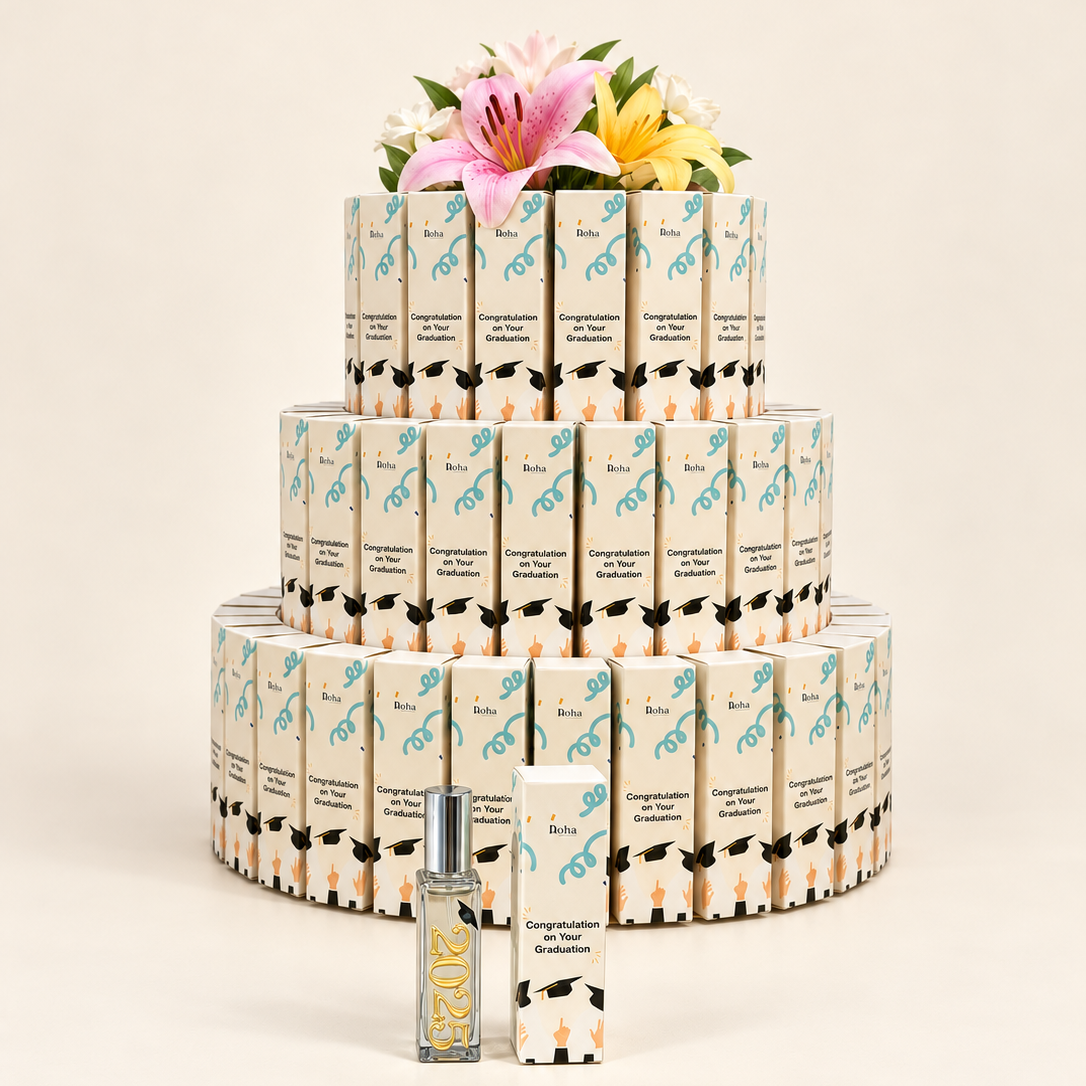 A tiered display of small rectangular cosmetic boxes wrapped in cream paper with black and teal graphics, topped with pink and yellow flowers. In front is a small clear bottle with a silver cap and a matching box, photographed on a warm off-white studio background.