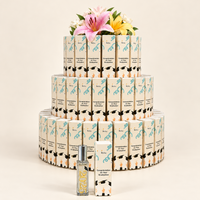 A tiered display of small rectangular cosmetic boxes wrapped in cream paper with black and teal graphics, topped with pink and yellow flowers. In front is a small clear bottle with a silver cap and a matching box, photographed on a warm off-white studio background.
