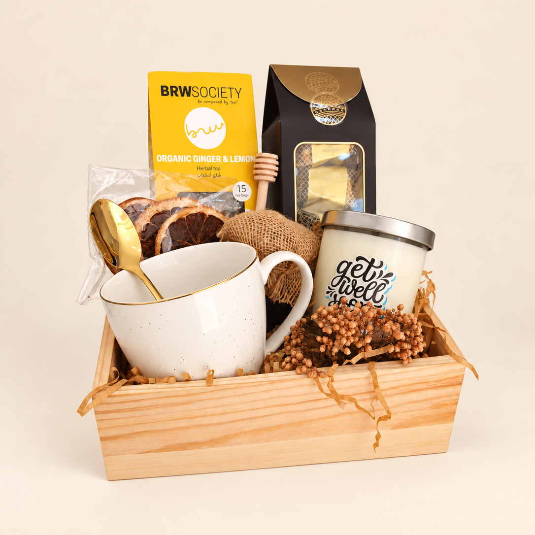 A curated gift basket in a light wood tray, featuring a white ceramic mug with gold trim and a gold spoon, a yellow BRW Society tea box, a black coffee bag, a white candle with blue lettering, and dried floral filler.