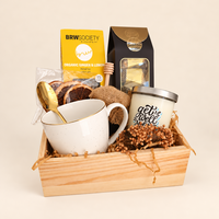 A curated gift basket in a light wood tray, featuring a white ceramic mug with gold trim and a gold spoon, a yellow BRW Society tea box, a black coffee bag, a white candle with blue lettering, and dried floral filler.