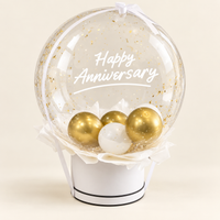A round clear balloon filled with gold confetti and the words 'Happy Anniversary' in white script, displayed on a white cylindrical stand with three metallic gold and one white ornament inside. The product is photographed against a clean, light beige background.
