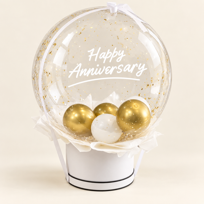 A round clear balloon filled with gold confetti and the words 'Happy Anniversary' in white script, displayed on a white cylindrical stand with three metallic gold and one white ornament inside. The product is photographed against a clean, light beige background.