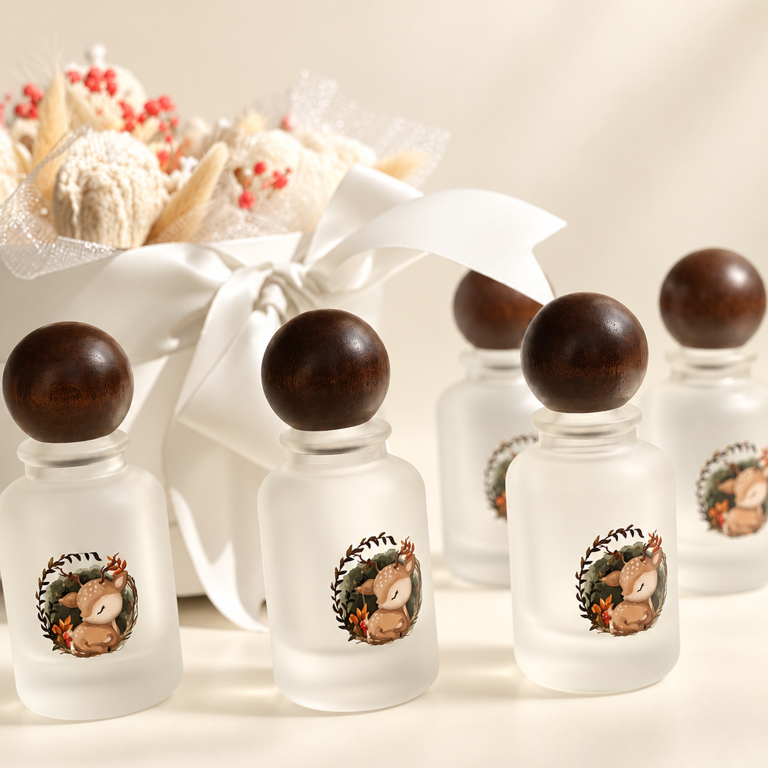 Four frosted glass perfume bottles with dark wooden caps and a printed deer illustration on the front, arranged on a light beige surface with a white ribbon and dried flowers in the background.