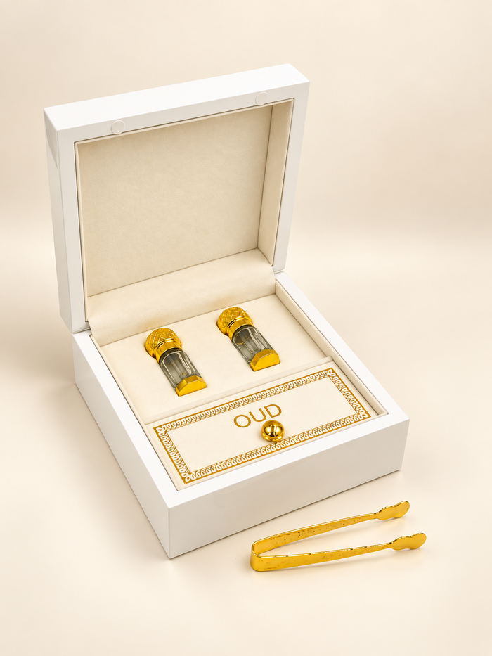 A pair of gold cufflinks with textured cylindrical tops, presented in an open white jewelry box with a cream velvet lining. A small gold tag inside the box reads 'OUD', and a pair of gold tongs rests outside the box on a warm beige background.