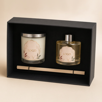 A black gift box containing a white candle in a glass jar with a silver lid, a clear glass reed diffuser bottle with a silver cap, and two wooden reeds, all labeled with generic 'LOGO' graphics, on a beige background.