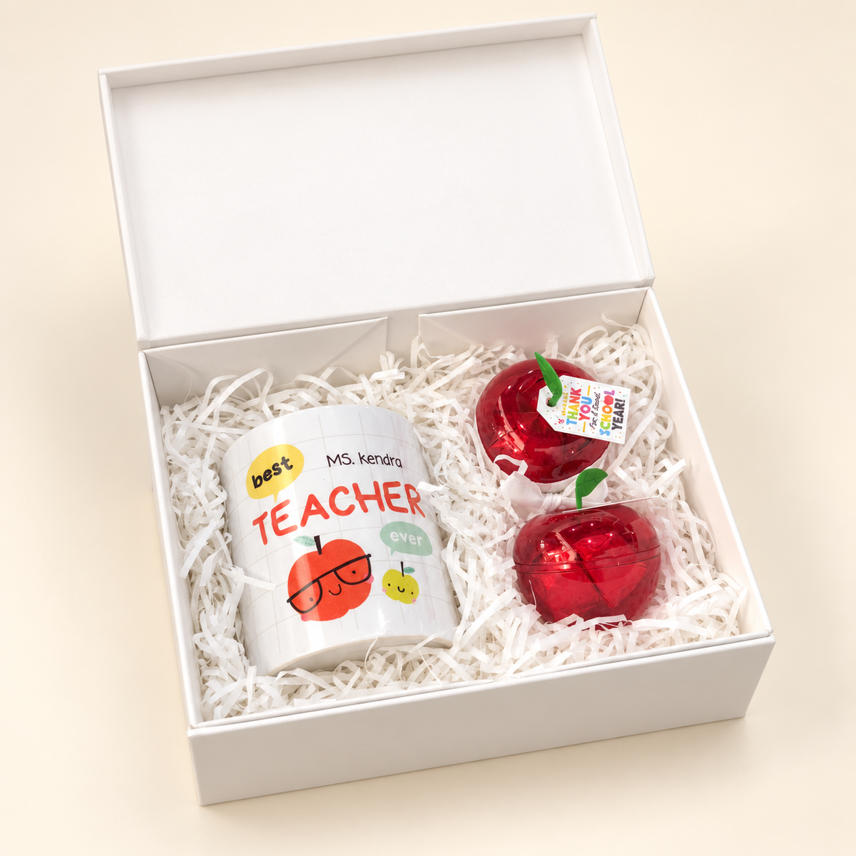 An open white gift box contains a printed teacher mug and two red apple-shaped items, one for chocolate and one for a message.