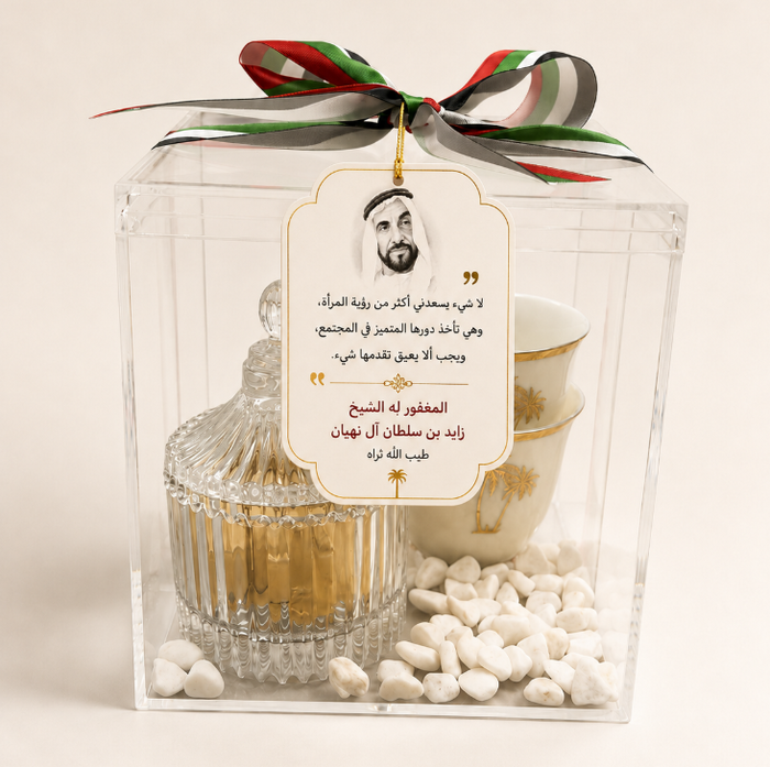 A clear acrylic gift box with a lid and a hanging tag, filled with a gold-toned decorative container, a white teacup with gold detailing, white stones, and a green, white, and red ribbon tied at the top. The tag features Arabic text and a portrait.