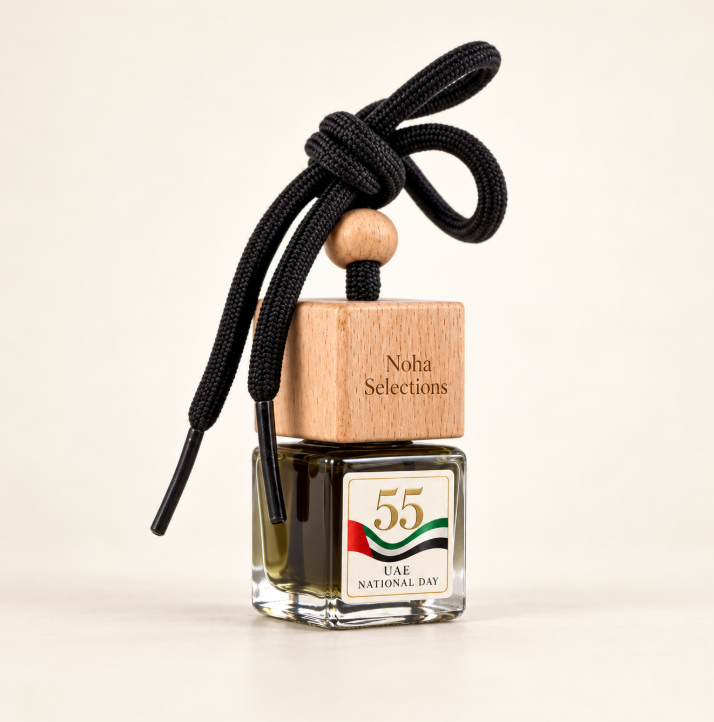 A glass bottle fragrance diffuser with a wooden cap engraved 'Noha Selections' and a black cord tied in a bow. The bottle has a UAE National Day label and is photographed on a clean cream background.