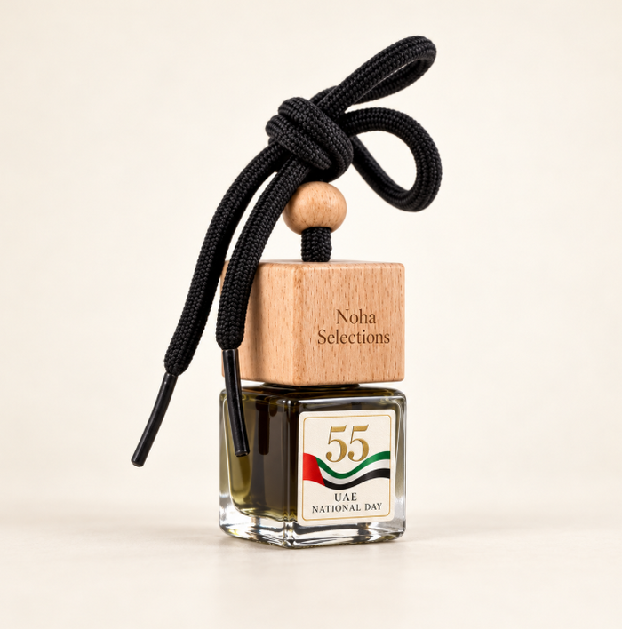 A glass bottle fragrance diffuser with a wooden cap engraved 'Noha Selections' and a black cord tied in a bow. The bottle has a UAE National Day label and is photographed on a clean cream background.