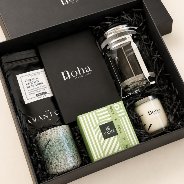 A curated gift box set from Noha Selection and Avantic, arranged in a black presentation box with black shredded paper. The set includes a green speckled ceramic candle, a green and white patterned soap box labeled Patchi, a black coffee bag, and a black book or notebook.