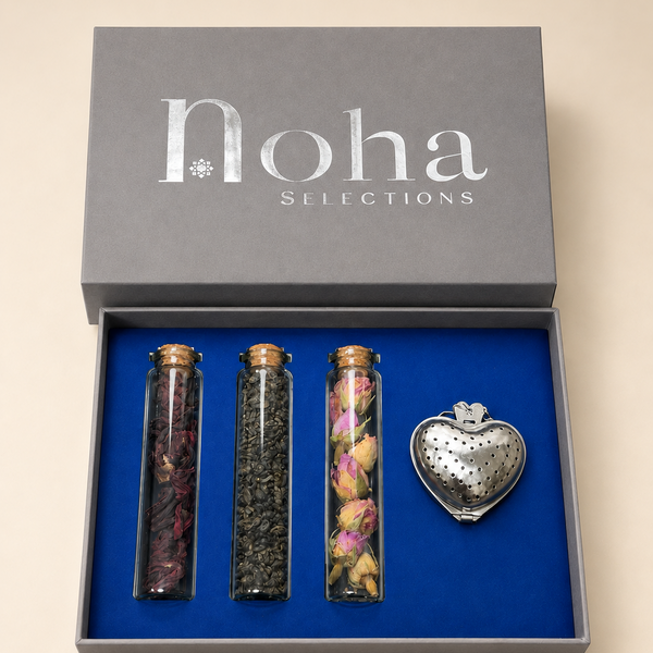 A Noha Selections gift set in a gray box with a blue interior, containing three glass vials with dried botanicals and a silver heart-shaped metal infuser.