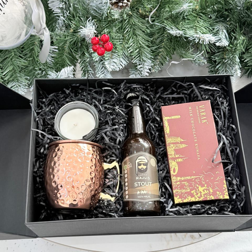 Gift set with a copper mug, bottle, and book in a box with Christmas decorations in the background.
