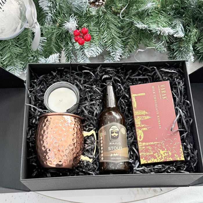 Gift set with a copper mug, bottle, and book in a box with Christmas decorations in the background.