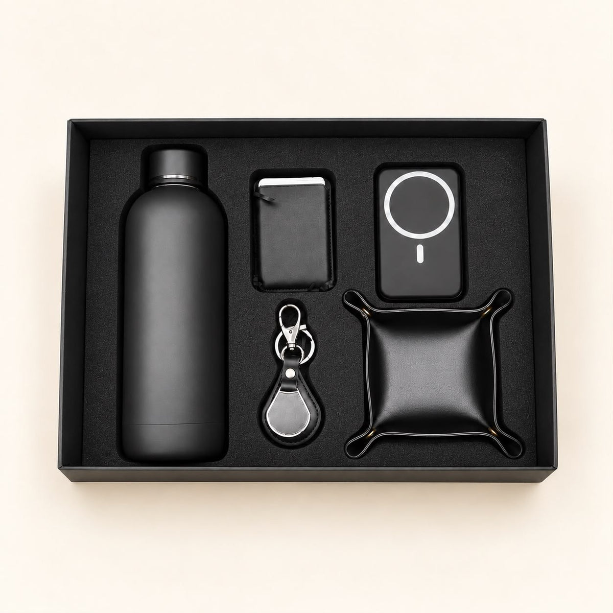 A black gift set containing a matte black bottle, a slim rectangular case, a circular magnetic charging pad, a keychain with a silver clasp, and a small black pouch, all arranged in a black presentation box on a light beige background.