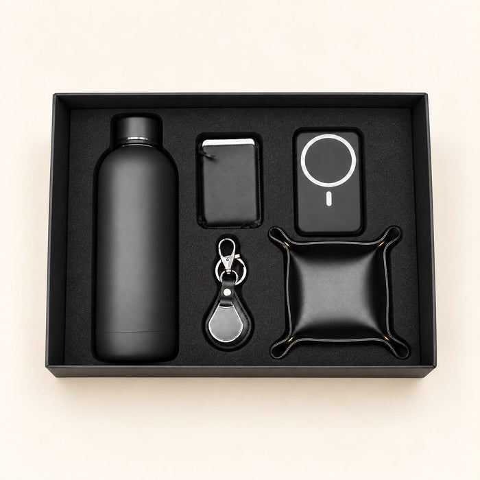 A black gift set containing a matte black bottle, a slim rectangular case, a circular magnetic charging pad, a keychain with a silver clasp, and a small black pouch, all arranged in a black presentation box on a light beige background.