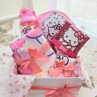 A pink Hello Kitty gift basket set on a marble table, containing a pink water bottle, a Hello Kitty pouch, a Hello Kitty card, and folded pink and white items tied with pink ribbons.