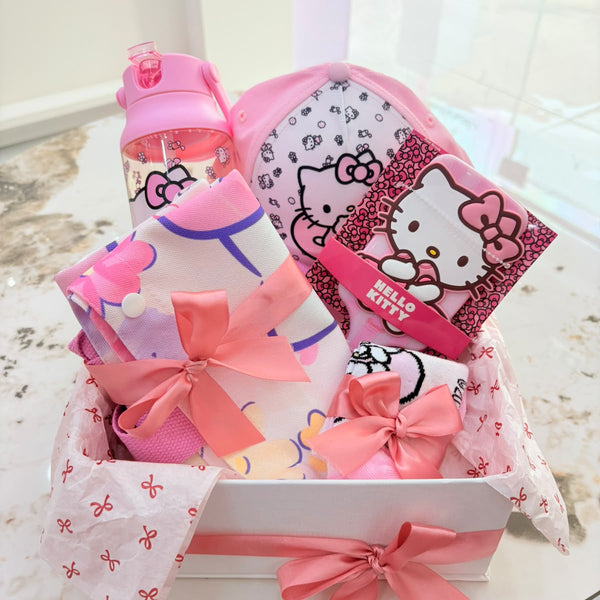 A pink Hello Kitty gift basket set on a marble table, containing a pink water bottle, a Hello Kitty pouch, a Hello Kitty card, and folded pink and white items tied with pink ribbons.