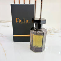 A bottle of Noha Selections perfume with golden-yellow liquid in a dark faceted glass bottle, shown next to its black packaging box with gold lettering on a light marble surface.
