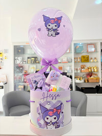A Sanrio-themed gift basket featuring a large lavender balloon with Kuromi, a white cylindrical box printed with Kuromi graphics and the name 'Hessa', and assorted pastel bath towels and packaged items inside, set in a bright retail store environment.