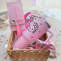 A Hello Kitty gift basket set in a woven wicker basket, containing a pink insulated tumbler with a Hello Kitty face, a pink hairbrush in its original packaging, a pink bow, and a pink patterned cloth, displayed on a marble surface.