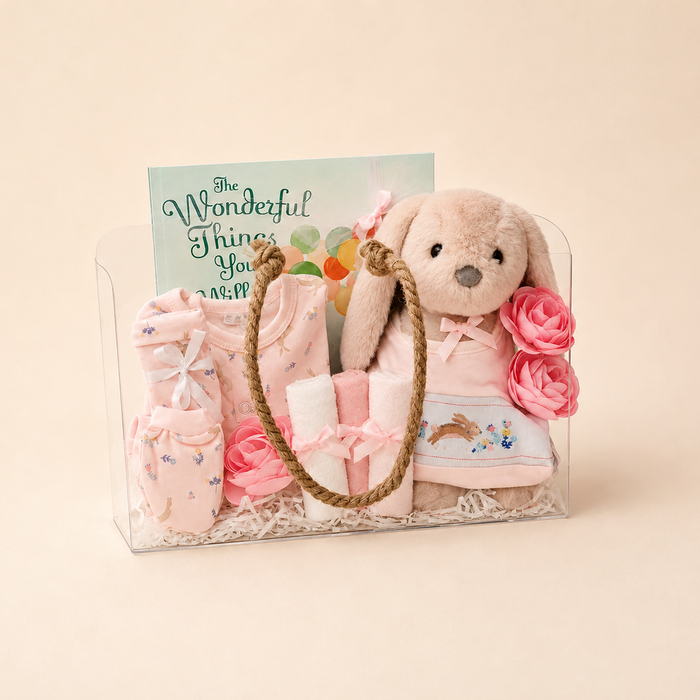 A baby gift basket set in a clear plastic case, featuring a plush bunny doll, pink baby clothes, a small plush hat, pink fabric roses, and a greeting card that reads 'The Wonderful Things You Will'.