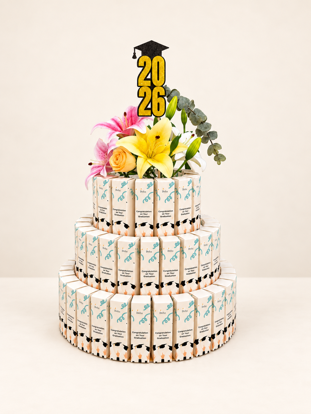 A three-tiered graduation cake made from wrapped candy bars, decorated with yellow and pink flowers and a '2026' graduation topper, set against a plain light beige background.