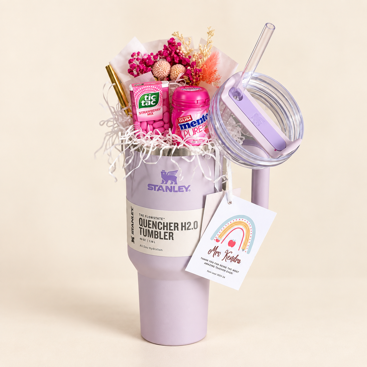 A lavender 40oz Stanley Quencher H2.0 tumbler filled with dried pink flowers, Tic Tac mints, Mentos gum, and a personalized teacher tag